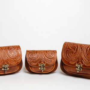 May include: Three brown leather saddlebags with intricate floral and swirl embossed designs. Each bag has a curved flap, a metal clasp, and a rounded bottom. The bags are in varying sizes, showcasing a cohesive collection.