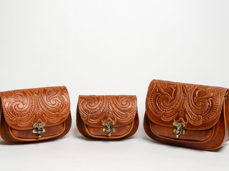 May include: Three brown leather saddlebags with intricate floral and swirl embossed designs. Each bag has a curved flap, a metal clasp, and a rounded bottom. The bags are in varying sizes, showcasing a cohesive collection.