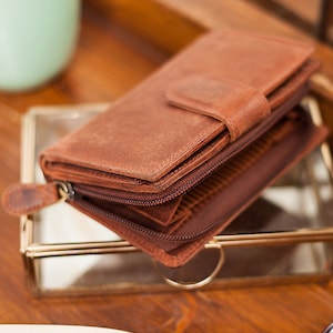 May include: A brown leather wallet with a zipper closure and a flap with a snap closure. The wallet is folded and sitting on a glass surface.