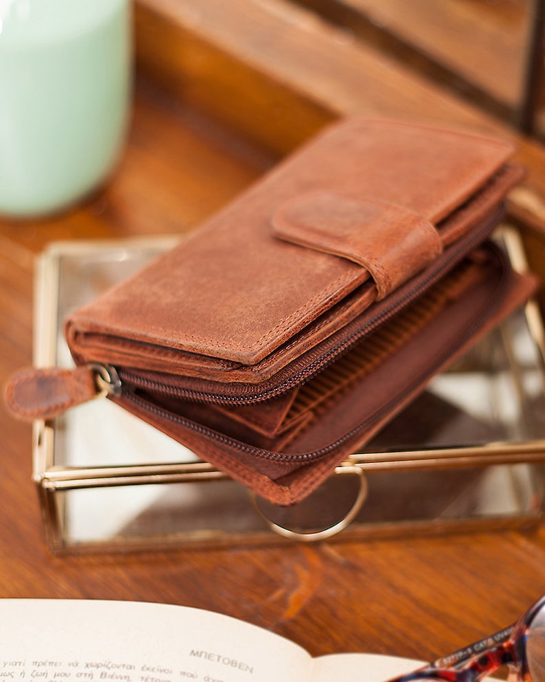 May include: A brown leather wallet with a zipper closure and a flap with a snap closure. The wallet is folded and sitting on a glass surface.