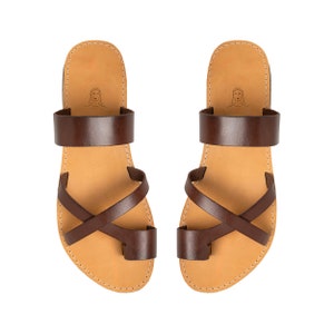 May include: A pair of dark brown leather sandals featuring a wide strap across the instep, a crisscross strap design over the midfoot, and a separate toe loop. The tan footbed includes a small line drawing of a face near the heel area.