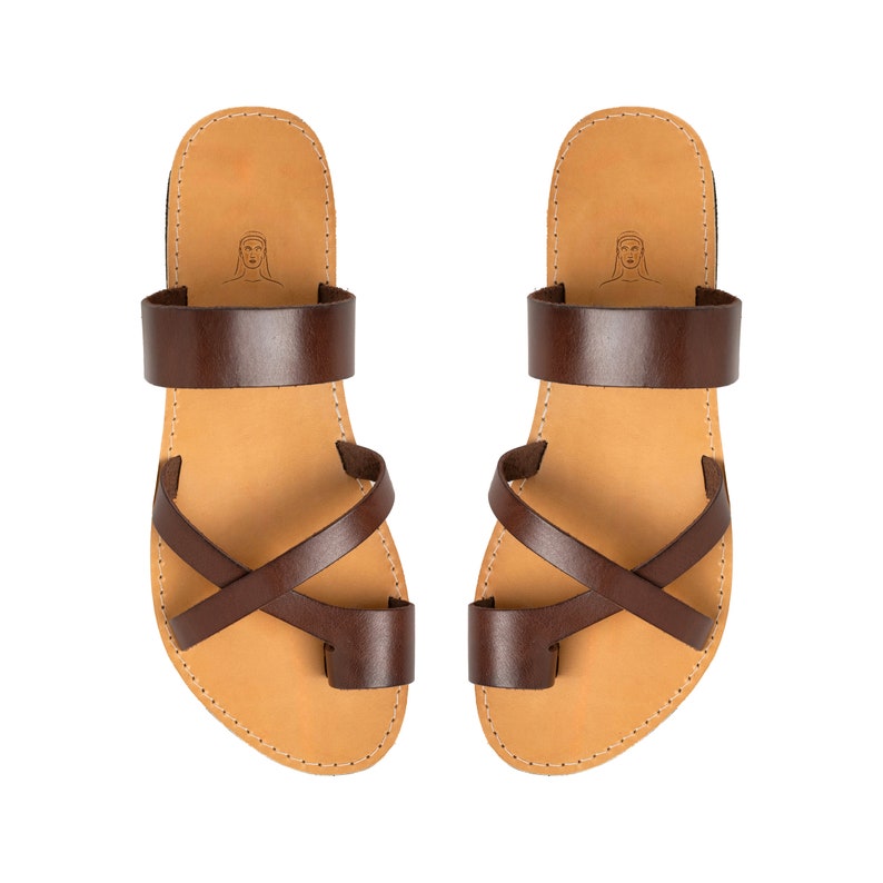 May include: A pair of dark brown leather sandals featuring a wide strap across the instep, a crisscross strap design over the midfoot, and a separate toe loop. The tan footbed includes a small line drawing of a face near the heel area.