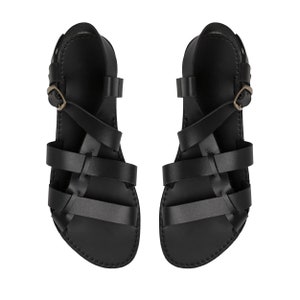 May include: A pair of black leather sandals featuring multiple horizontal straps across the foot, a secure ankle strap with a metallic buckle, and a flat sole. The design includes a series of interconnected straps that wrap around the foot.