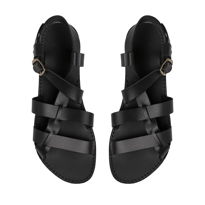 May include: A pair of black leather sandals featuring multiple horizontal straps across the foot, a secure ankle strap with a metallic buckle, and a flat sole. The design includes a series of interconnected straps that wrap around the foot.