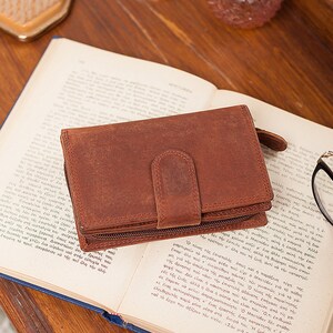 May include: A brown leather wallet with a zipper closure and a flap with a snap closure. The wallet is lying open on a book.