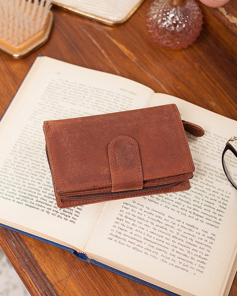 May include: A brown leather wallet with a zipper closure and a flap with a snap closure. The wallet is lying open on a book.