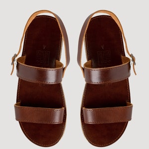 May include: A pair of dark brown leather sandals featuring two horizontal straps across the foot, a heel strap with a small metal buckle, and a flat sole. The footbed displays a stamped size 4 and a small icon indicating leather construction.
