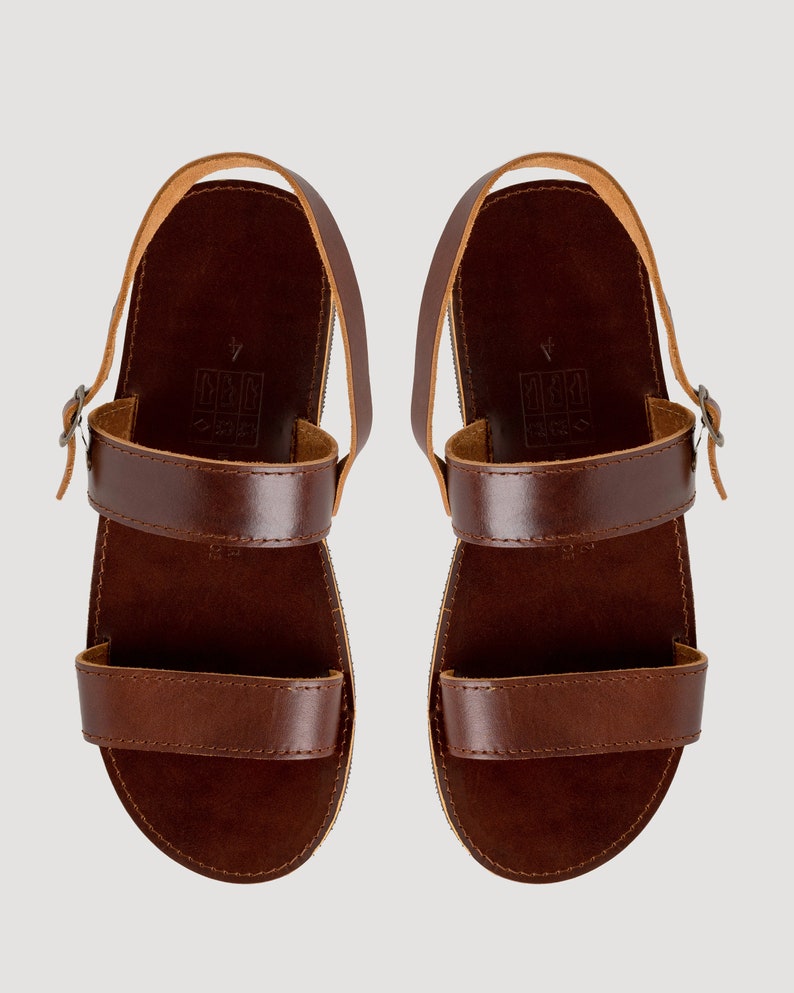 May include: A pair of dark brown leather sandals featuring two horizontal straps across the foot, a heel strap with a small metal buckle, and a flat sole. The footbed displays a stamped size 4 and a small icon indicating leather construction.