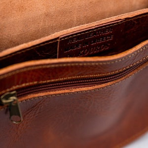 May include: The inside of a brown leather bag with a zipper closure. The bag is lined with brown leather and has a label that reads "Real Leather Made in Greece by Kouros".