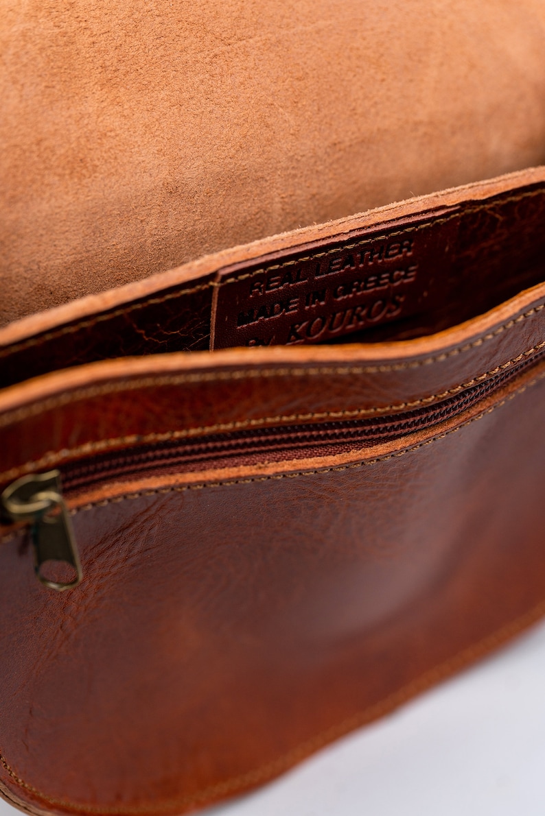 May include: The inside of a brown leather bag with a zipper closure. The bag is lined with brown leather and has a label that reads "Real Leather Made in Greece by Kouros".