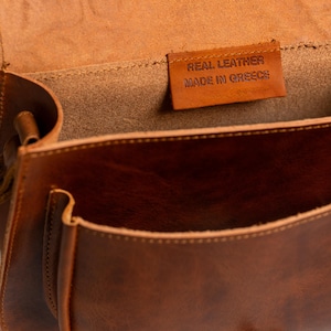 May include: Open view of a brown leather satchel bag. The bag has a front pocket and a flap closure. Inside the bag, a small leather tag reads "REAL LEATHER MADE IN GREECE". The bag is made of a rich, warm brown leather.