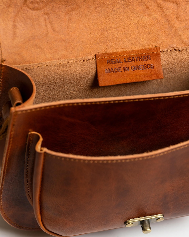 May include: Open view of a brown leather satchel bag. The bag has a front pocket and a flap closure. Inside the bag, a small leather tag reads "REAL LEATHER MADE IN GREECE". The bag is made of a rich, warm brown leather.