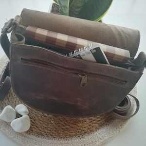 Puede incluir: Un bolso de cuero marr&oacute;n con un bolsillo con cremallera y cierre de solapa. El bolso est&aacute; abierto y contiene un cuaderno y una libreta de espiral.