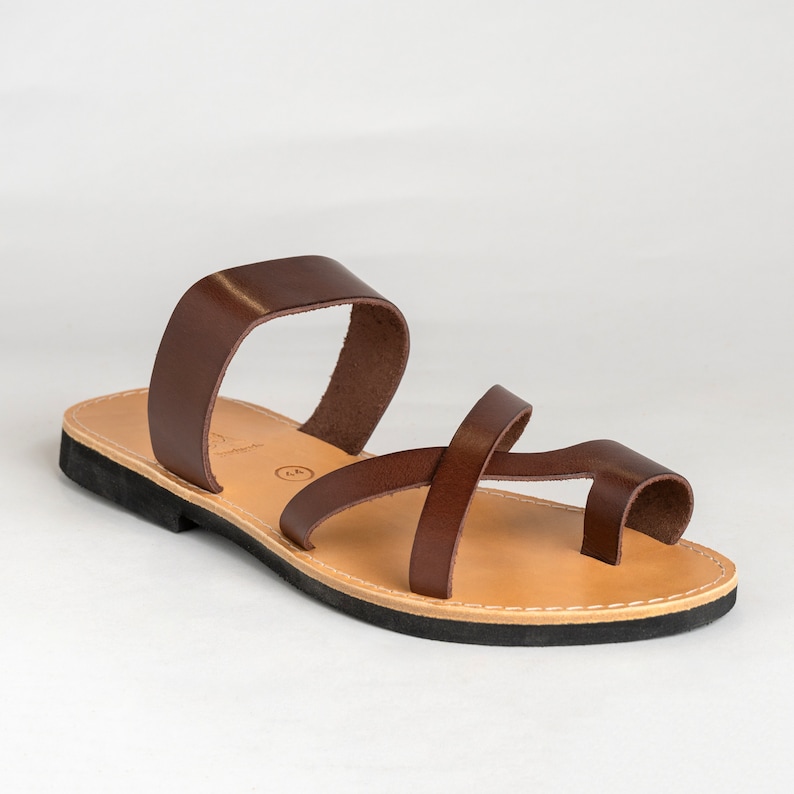 May include: A dark brown leather sandal featuring a wide strap across the midfoot and a thinner strap that crosses over the top of the foot, looping around the big toe. The sandal has a tan leather insole and a black rubber outsole.
