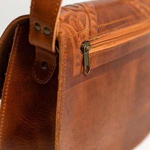 May include: Close-up of a brown leather shoulder bag with a flap closure and a zipper. The bag features embossed floral detailing on the top and a long adjustable strap. The hardware is a brass color.