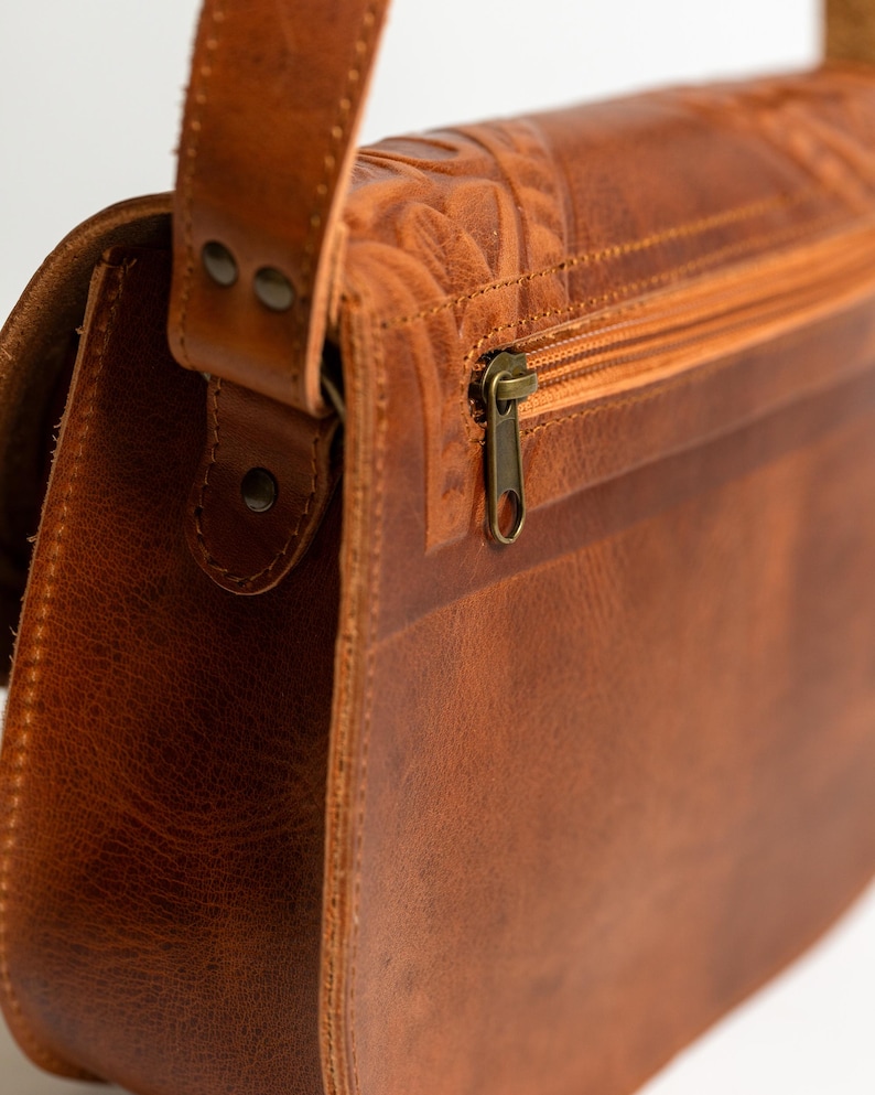 May include: Close-up of a brown leather shoulder bag with a flap closure and a zipper. The bag features embossed floral detailing on the top and a long adjustable strap. The hardware is a brass color.