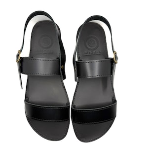 May include: A pair of black leather sandals featuring a wide toe strap, a mid-foot strap with a brass-toned metal buckle, and a heel strap. The footbed is embossed with the text HITONAS and a circular logo design. The sandals have a smooth finish.