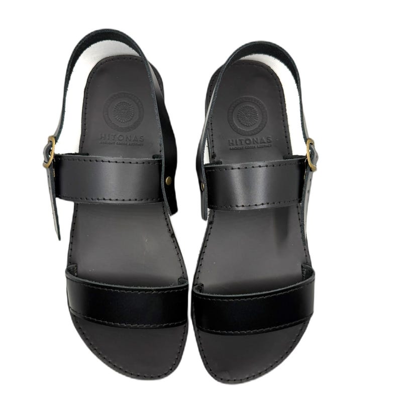 May include: A pair of black leather sandals featuring a wide toe strap, a mid-foot strap with a brass-toned metal buckle, and a heel strap. The footbed is embossed with the text HITONAS and a circular logo design. The sandals have a smooth finish.