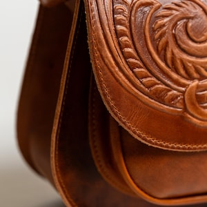 May include: Close-up of a brown leather satchel with intricate floral and scrollwork carving on the flap. The bag features a curved shape and a stitched design, showcasing a vintage aesthetic.