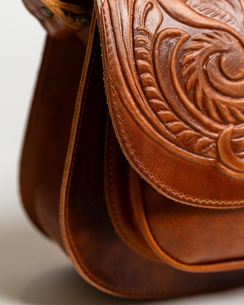 May include: Close-up of a brown leather satchel with intricate floral and scrollwork carving on the flap. The bag features a curved shape and a stitched design, showcasing a vintage aesthetic.