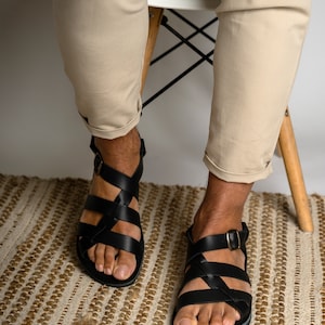 May include: A pair of black leather sandals featuring multiple crisscrossing straps across the top of the foot and an adjustable ankle strap with a small metal buckle. The sandals have a flat sole and an open-toe design, providing a secure fit.