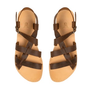 May include: A pair of brown leather sandals featuring multiple crisscrossing straps, an adjustable ankle strap with a metal buckle, and a light tan footbed. A small line drawing of a person's head is printed on the heel area of each sandal.