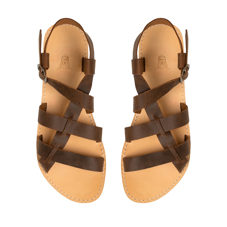 May include: A pair of brown leather sandals featuring multiple crisscrossing straps, an adjustable ankle strap with a metal buckle, and a light tan footbed. A small line drawing of a person's head is printed on the heel area of each sandal.