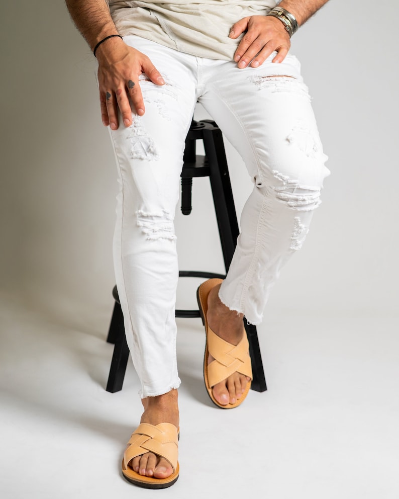 May include: White denim jeans featuring distressed rips and frayed hems, paired with light tan leather slide sandals with a woven crisscross strap design. The jeans have a slim fit and a classic five-pocket construction with metal rivets.
