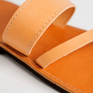 May include: A tan leather sandal featuring a wide upper strap with white stitching and a thinner diagonal strap across the footbed. The sandal has a smooth tan leather insole and a dark brown textured sole, showcasing clean construction details.