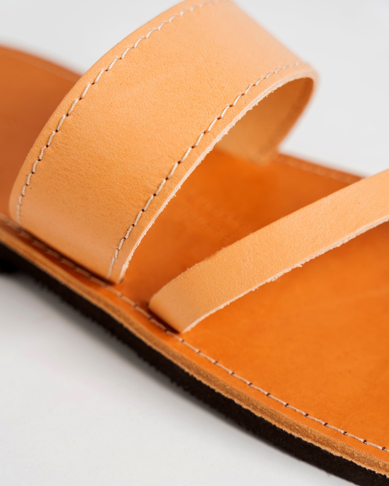 May include: A tan leather sandal featuring a wide upper strap with white stitching and a thinner diagonal strap across the footbed. The sandal has a smooth tan leather insole and a dark brown textured sole, showcasing clean construction details.