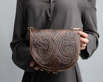 May include: A brown leather satchel bag with intricate floral carvings. The bag has a flap closure and a strap.