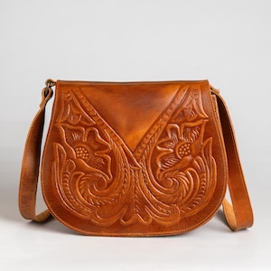 May include: A brown leather crossbody bag with a floral embossed design. The bag has a long adjustable strap and a flap closure. The intricate carving showcases detailed flowers and leaves, adding a rustic and artistic touch to the accessory.