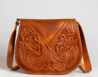 May include: A brown leather crossbody bag with a floral embossed design. The bag features a long adjustable strap and a flap closure. The intricate carving showcases detailed flowers and leaves, adding a rustic and artistic touch to the accessory.