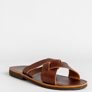 May include: A dark brown leather slide sandal featuring two wide straps that cross over the top of the foot. The sandal has a flat, dark brown sole with a visible light tan edge along the perimeter of the footbed and the strap construction.