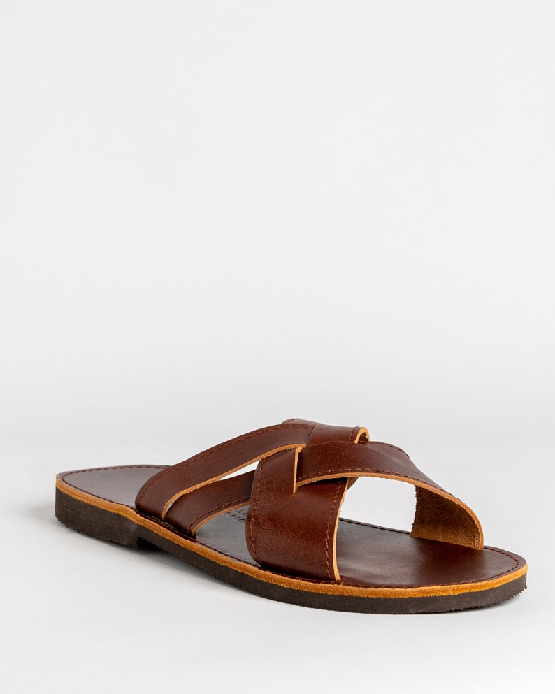 May include: A dark brown leather slide sandal featuring two wide straps that cross over the top of the foot. The sandal has a flat, dark brown sole with a visible light tan edge along the perimeter of the footbed and the strap construction.