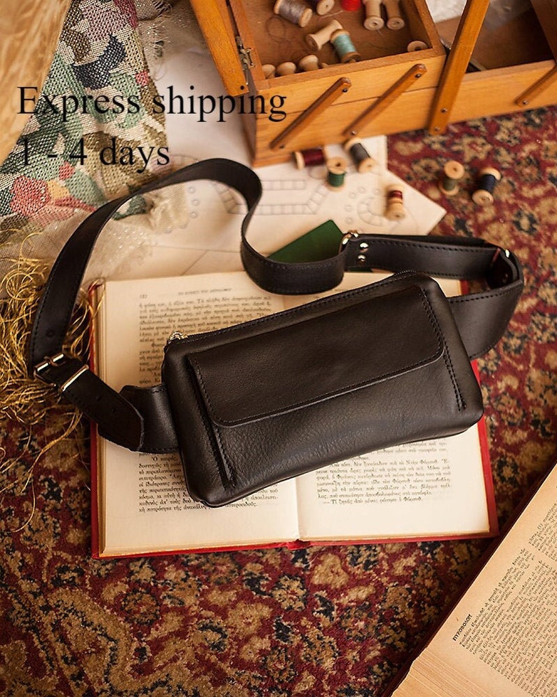 May include: Black leather fanny pack with a flap closure and adjustable strap. The bag is lying on an open book with red edges. The background includes a wooden sewing box and spools of thread.