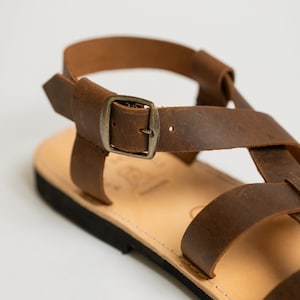 May include: A brown leather sandal featuring a flat sole, a light tan footbed, and a series of crisscrossing straps secured by a metallic buckle. The design includes a heel strap and a black outsole, showcasing a simple, functional construction.