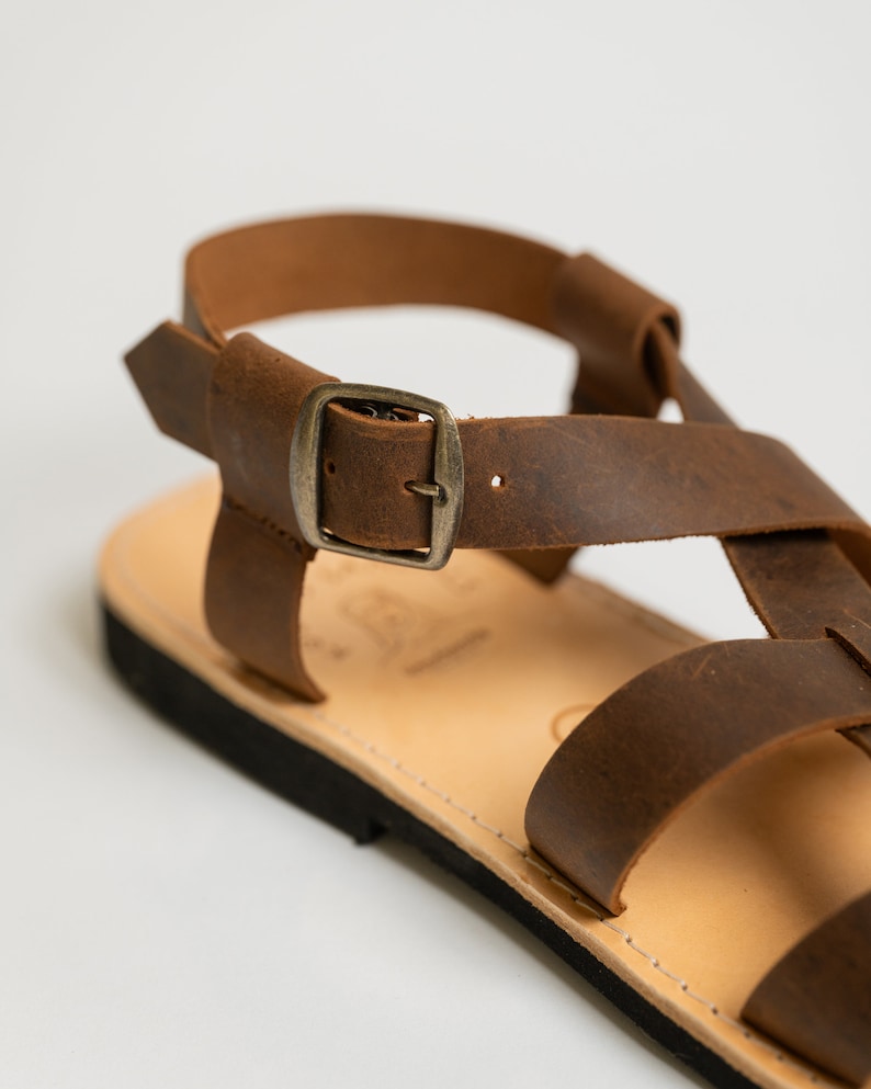 May include: A brown leather sandal featuring a flat sole, a light tan footbed, and a series of crisscrossing straps secured by a metallic buckle. The design includes a heel strap and a black outsole, showcasing a simple, functional construction.