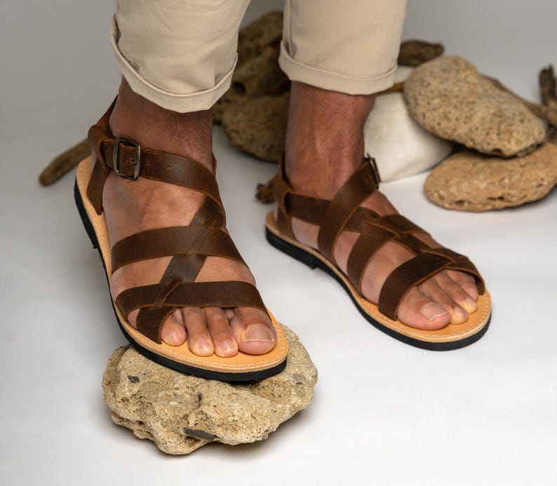 May include: A pair of brown leather sandals featuring multiple crisscrossing straps across the top of the foot, an adjustable ankle strap with a dark metal buckle, a light tan footbed, and a thin black sole, worn with light beige trousers.