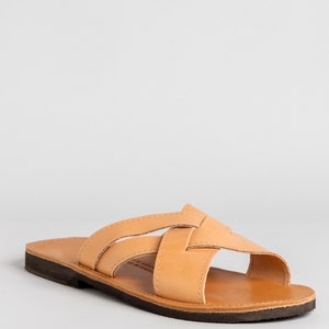 May include: A tan leather slide sandal featuring a crisscross strap design across the top. The sandal has a smooth, light brown leather footbed and a dark brown, flat rubber sole. The construction includes visible stitching along the straps.