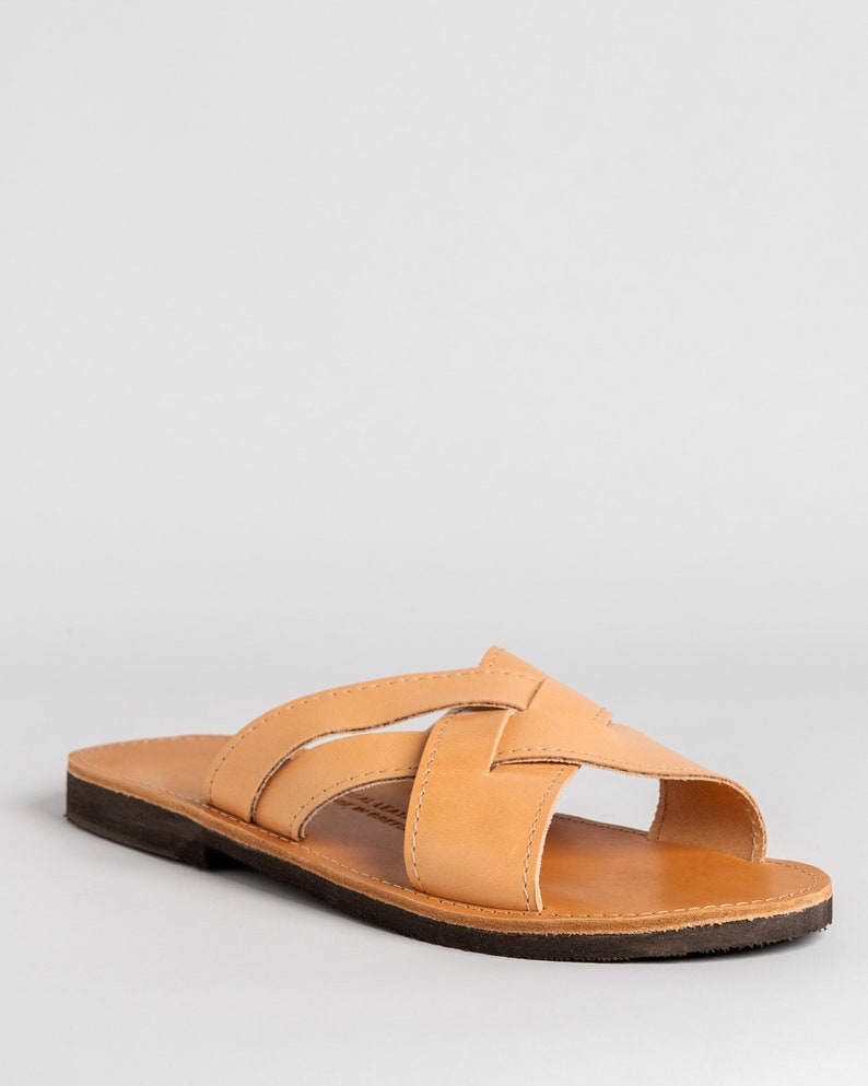 May include: A tan leather slide sandal featuring a crisscross strap design across the top. The sandal has a smooth, light brown leather footbed and a dark brown, flat rubber sole. The construction includes visible stitching along the straps.