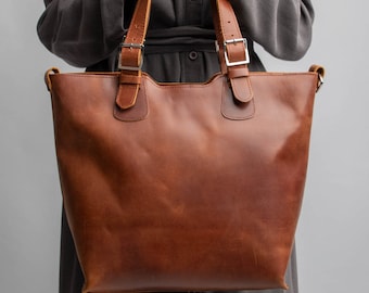 May include: A brown leather tote bag with two straps and silver buckles. The bag is held by a person wearing a gray shirt.