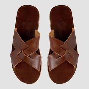 May include: A pair of brown leather sandals featuring a crisscross strap design across the top. The footbed is dark brown with a visible size 4 imprint and a square icon graphic. The edges of the straps show a lighter tan leather interior.