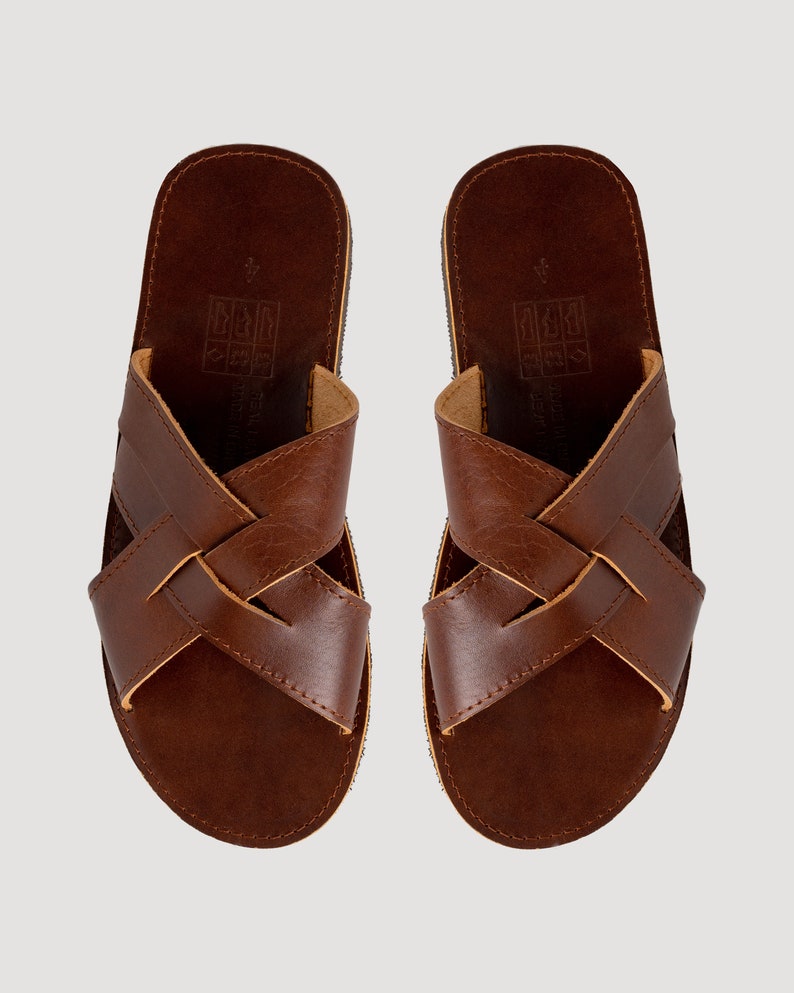 May include: A pair of brown leather sandals featuring a crisscross strap design across the top. The footbed is dark brown with a visible size 4 imprint and a square icon graphic. The edges of the straps show a lighter tan leather interior.
