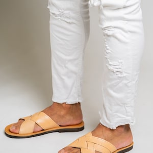 May include: A pair of light tan leather slide sandals with a woven crisscross strap design and a dark brown sole, worn with white distressed denim jeans featuring frayed hems and ripped details at the knees and shins.
