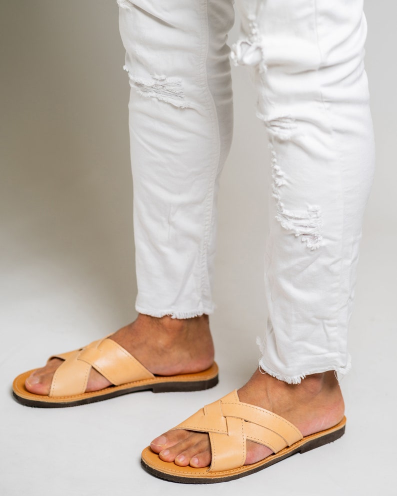 May include: A pair of light tan leather slide sandals with a woven crisscross strap design and a dark brown sole, worn with white distressed denim jeans featuring frayed hems and ripped details at the knees and shins.