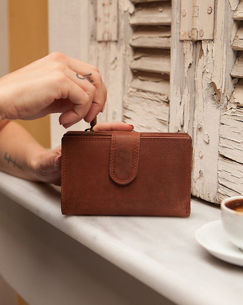 May include: A brown leather wallet with a flap closure and a metal zip. The wallet is sitting on a white surface.