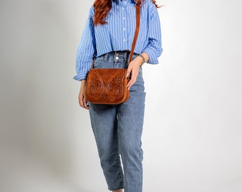 May include: A woman wearing a blue striped shirt and blue jeans is carrying a brown leather crossbody bag with intricate floral designs.
