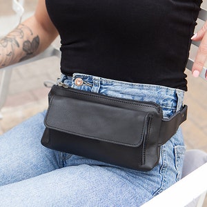 May include: A black leather fanny pack with a rectangular design and a flap closure. The pack is worn around the waist over blue jeans. The fanny pack has a zipper closure and a black adjustable belt.