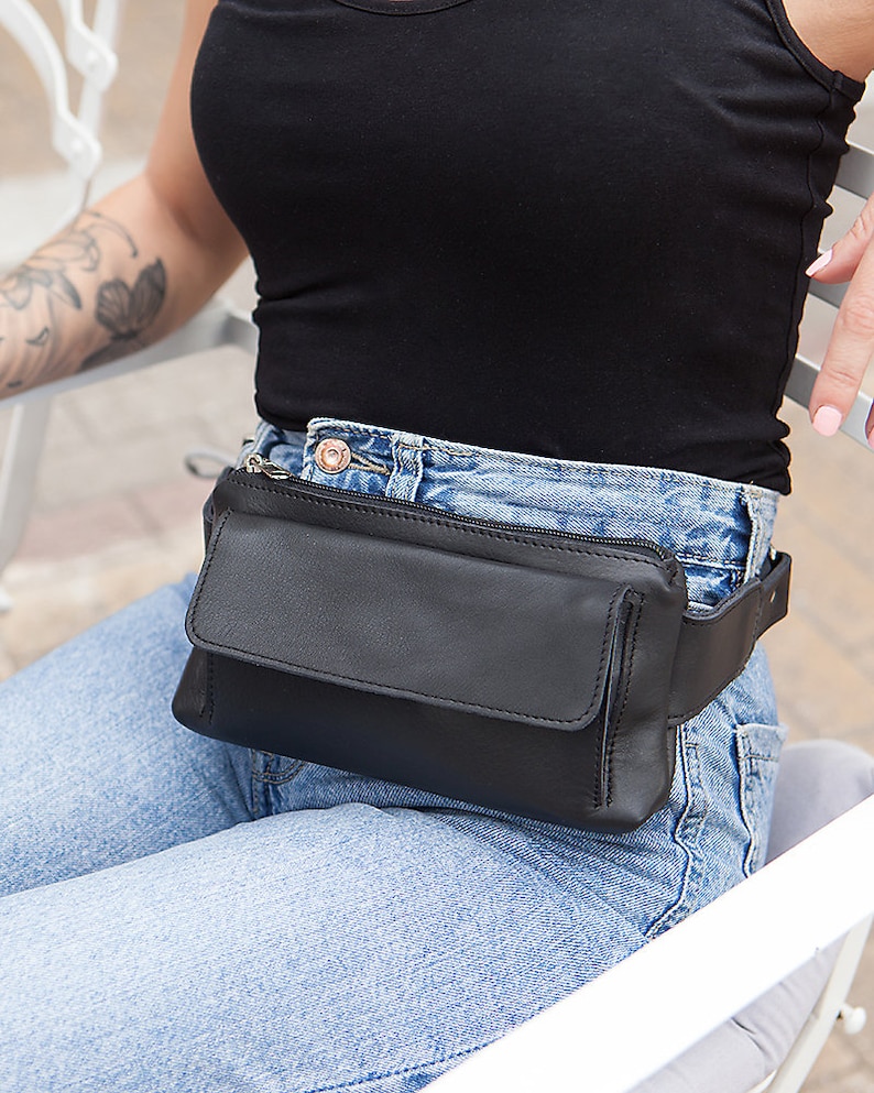 May include: A black leather fanny pack with a rectangular design and a flap closure. The pack is worn around the waist over blue jeans. The fanny pack has a zipper closure and a black adjustable belt.
